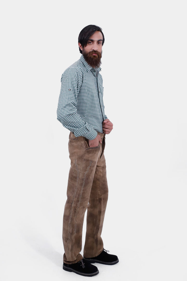 The man is standing at a slight angle, showing the full length of the Addison Oktoberfest Lederhosen Long and the green checkered shirt. He has one hand in his pocket and looks towards the camera.