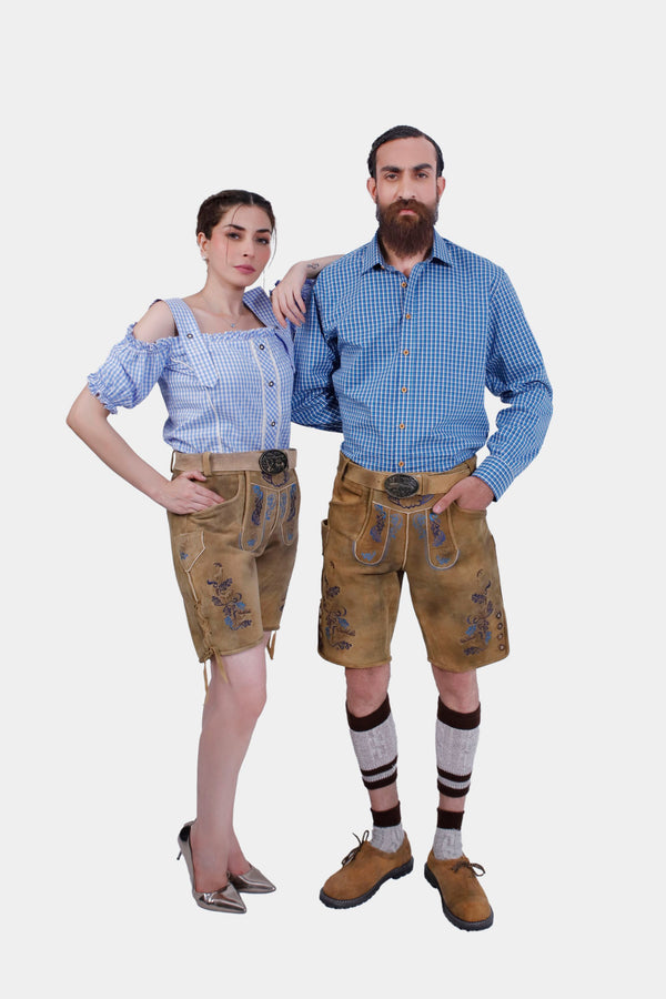 A couple wearing the Adirondack Couple Outfit. The man is dressed in a blue and white gingham check shirt with brown leather lederhosen featuring blue floral embroidery. The woman is wearing a matching blue and white gingham check blouse with brown leather lederhosen. Trachtenmode für Paare