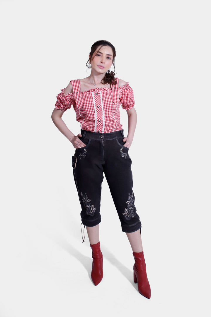 Full view of a woman wearing a red and white checkered off-shoulder blouse with ruffled edges, paired with black lederhosen and red boots, standing confidently with hands in pockets. damen lederhose schwarz tracht ,  lederhose damen tracht schwarz