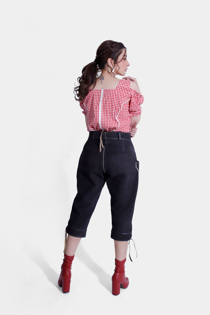 Back view of a woman wearing a red and white checkered off-shoulder blouse with ruffled edges and black lederhosen, standing with a relaxed pose