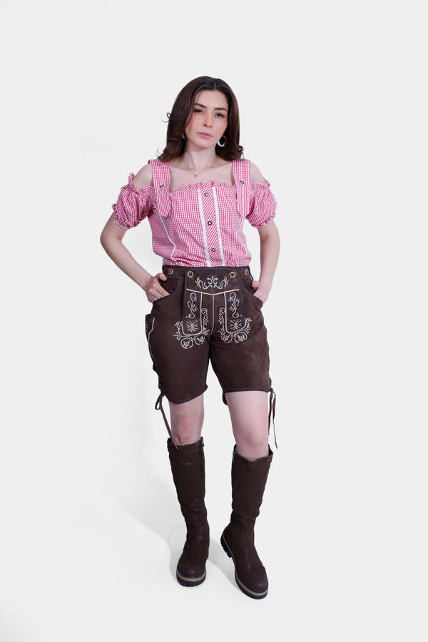 Woman wearing a traditional Bavarian outfit featuring light brown knee-length lederhosen and a red and white checkered off-shoulder shirt with ruffled sleeves. damen lederhose tracht​ , lederhose tracht damen​