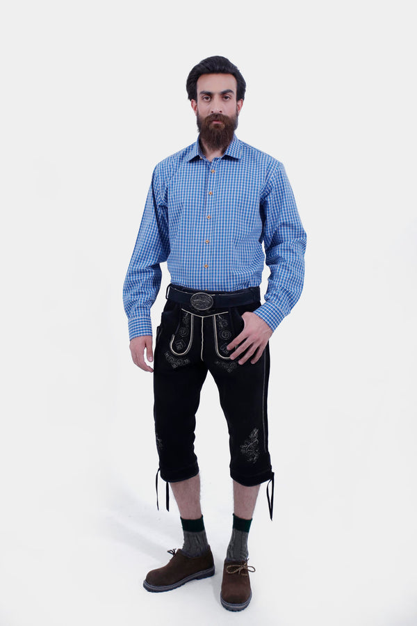 A man with a beard wearing a blue checkered shirt and black Bodensee Bundhosen with traditional socks and brown shoes, standing with his hands by his sides.