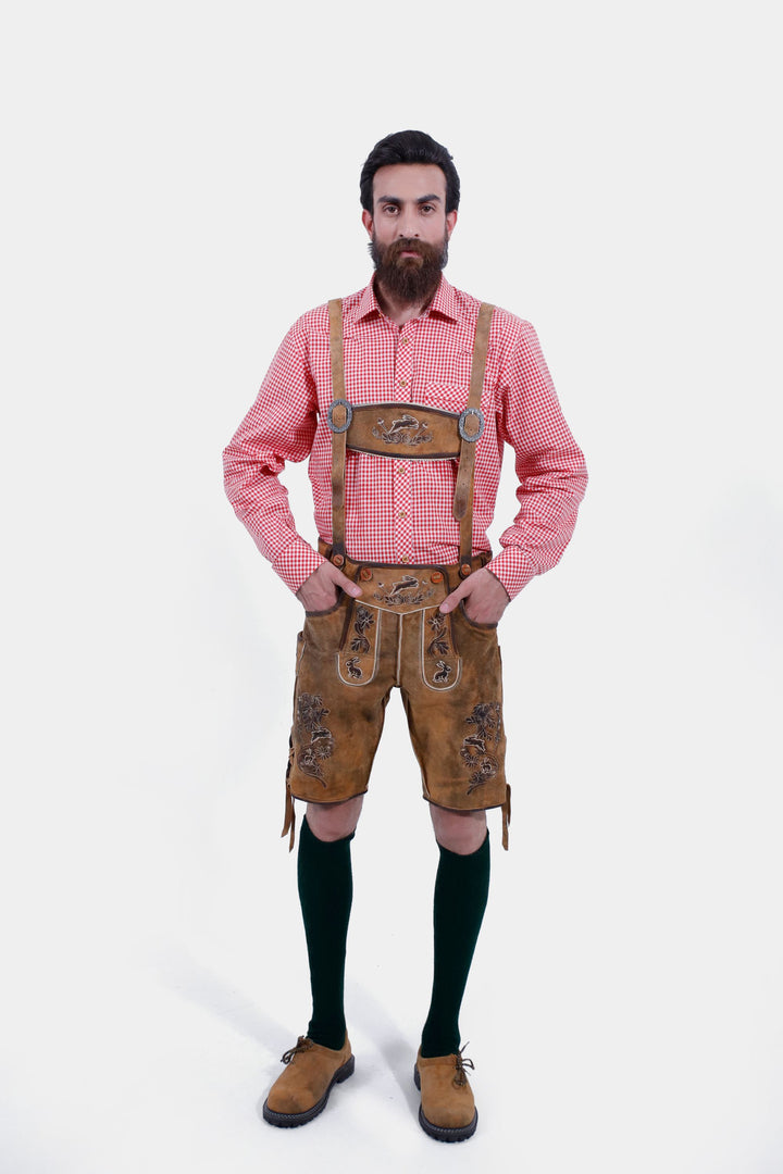 Full-body view of a man wearing Chiemsee Lederhosen with suspenders and a pink checkered shirt, standing with hands in pockets.