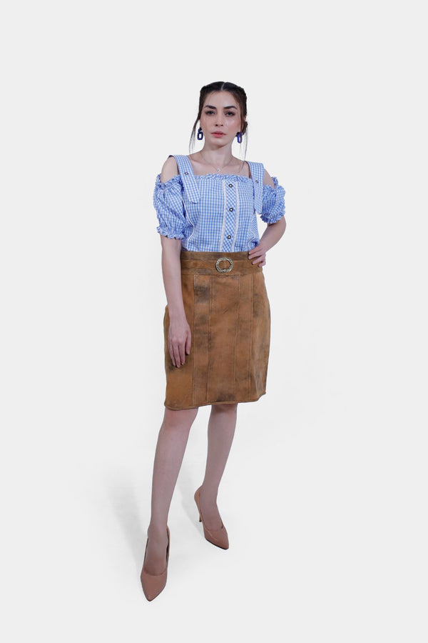 Close-up view of a Clara Lederhosen skirt, showcasing the detailed texture of the brown leather material and the decorative belt buckle, with the woman crossing her arms over her blue and white checkered blouse.