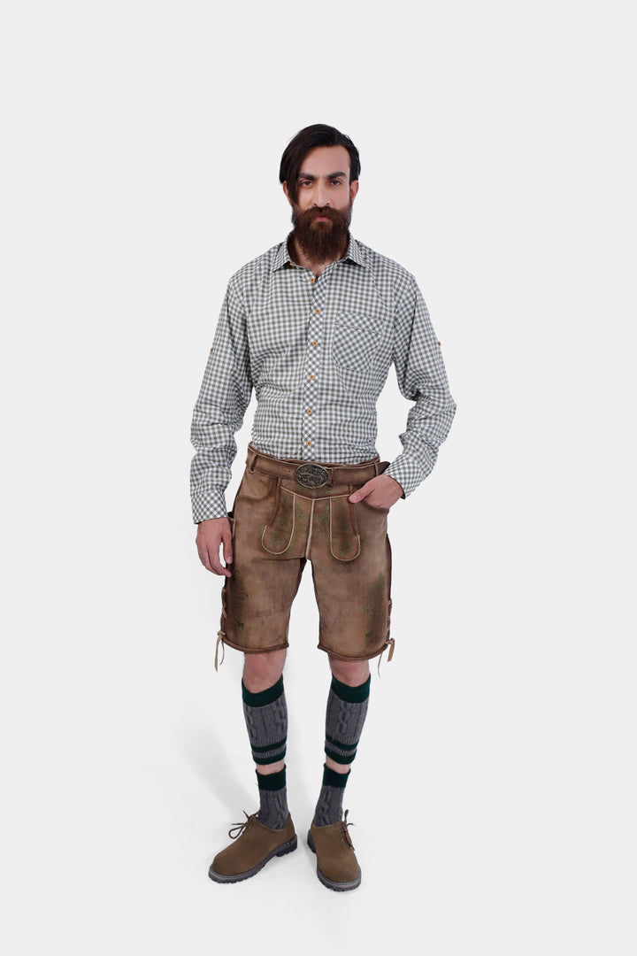 Full body view of a man wearing a green and white checkered shirt and brown leather lederhosen with knee-high socks and brown shoes, standing with a serious expression. lederhose braun herren​, lederhose herren braun