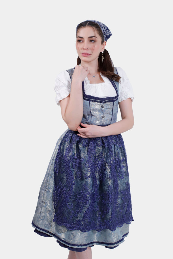 Woman in a Dirndl Maria Lara, featuring a laced bodice and floral-patterned apron, posing thoughtfully with hand on chin.