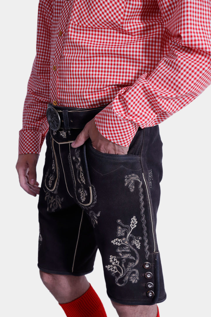 Side view of the Everglades Lederhosen Outfit, focusing on the detailed white embroidery on the black leather lederhosen, paired with a red and white checkered shirt and hand in the pocket.