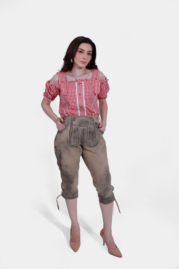 Woman in a red and white checkered off-shoulder blouse and light brown leather lederhosen, standing with hands in pockets, showing front view. lederhosen damen tracht​ , damen trachten lederhose knielang​