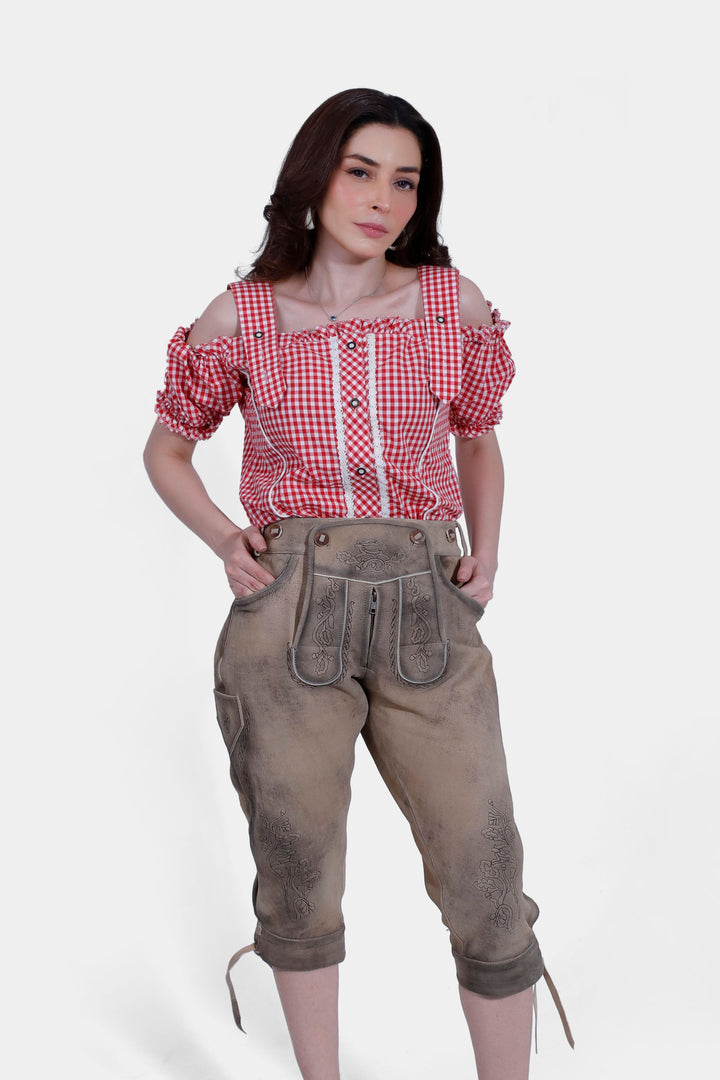 Woman in red and white checkered off-shoulder blouse and light brown lederhosen, posing with hands in pockets, displaying a close-up front view.