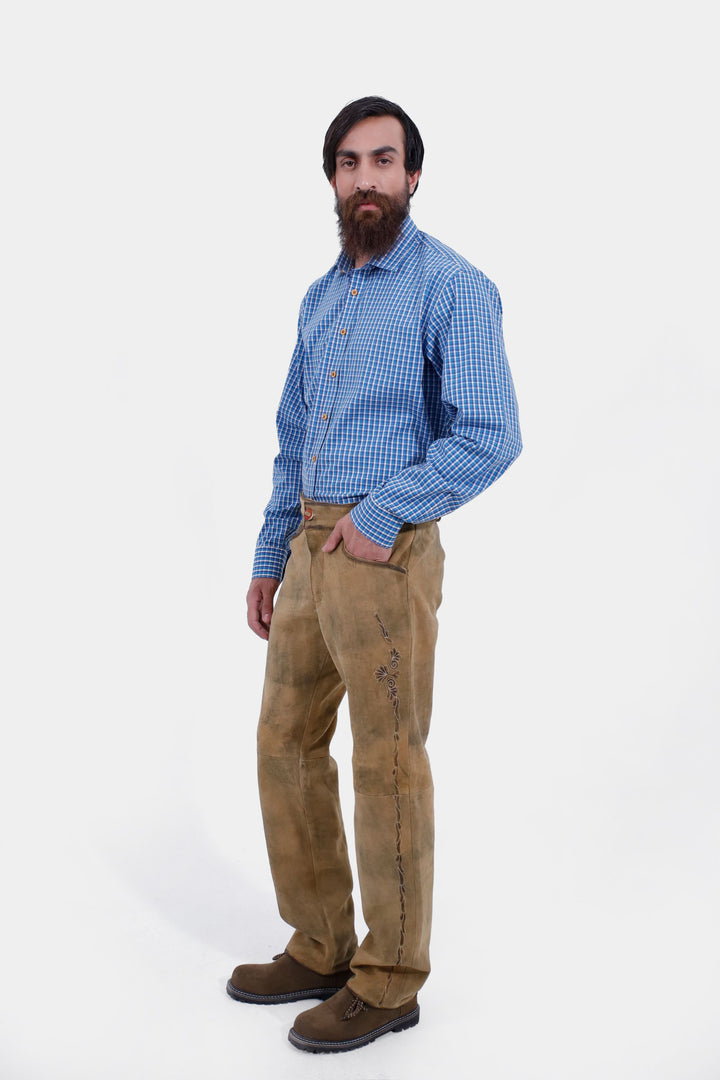 The man standing in a side profile, wearing the Goretzka lederhosen and blue checkered shirt, highlighting the overall fit and design of the lederhosen.