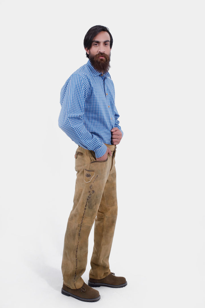 The man is standing at a slight angle, showing the full length of the Goretzka lederhosen and the blue checkered shirt. He has one hand in his pocket and looks towards the camera.