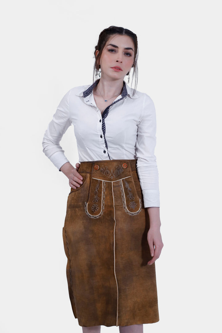 Close-up front view of a woman wearing the Griessee Lederhosen Skirt in brown leather paired with a white button-up shirt, focusing on the skirt's embroidery, buttons, and texture.