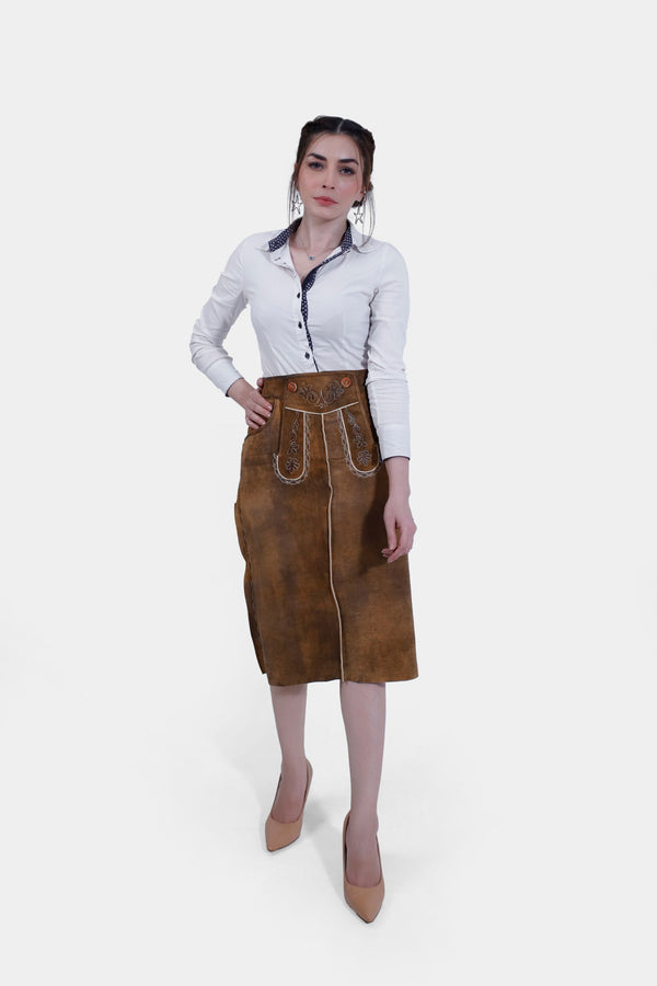 Woman wearing the Griessee Lederhosen Skirt in brown leather paired with a white button-up shirt, standing with one hand on her hip and the other by her side.