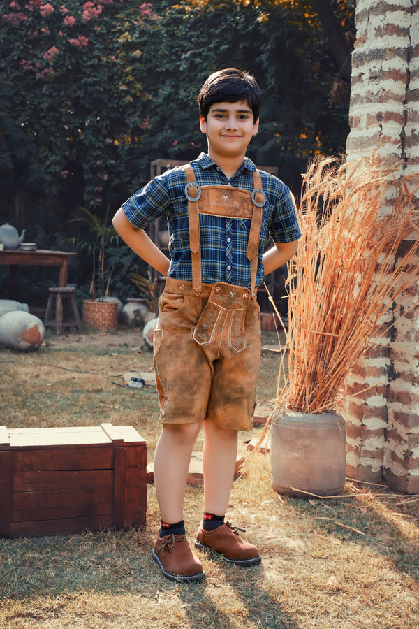 A young boy stands outdoors wearing Jakob Jäger Lederhosen, featuring a rustic amber color and traditional Bavarian design, with suspenders and detailed embroidery. He pairs the lederhosen with a blue plaid shirt and brown shoes, posing confidently with hands on his hips in a charming, rustic garden setting.