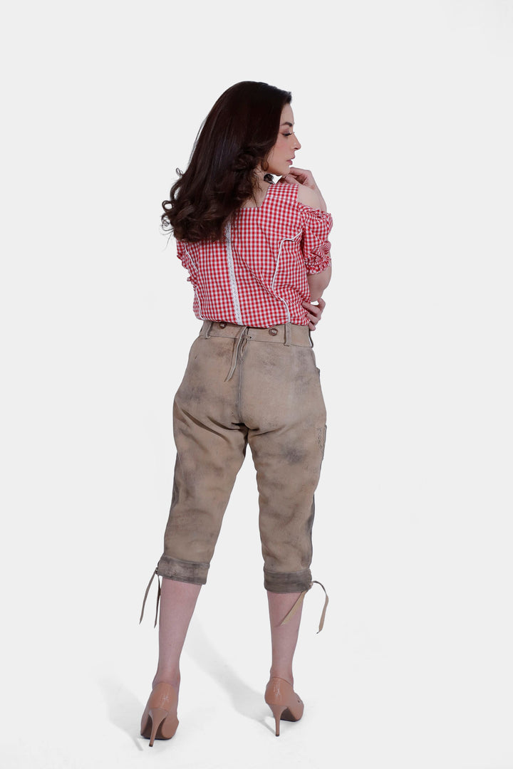 Full-body view of the woman in a red checkered blouse and brown lederhosen, standing with her legs crossed and looking directly at the camera.