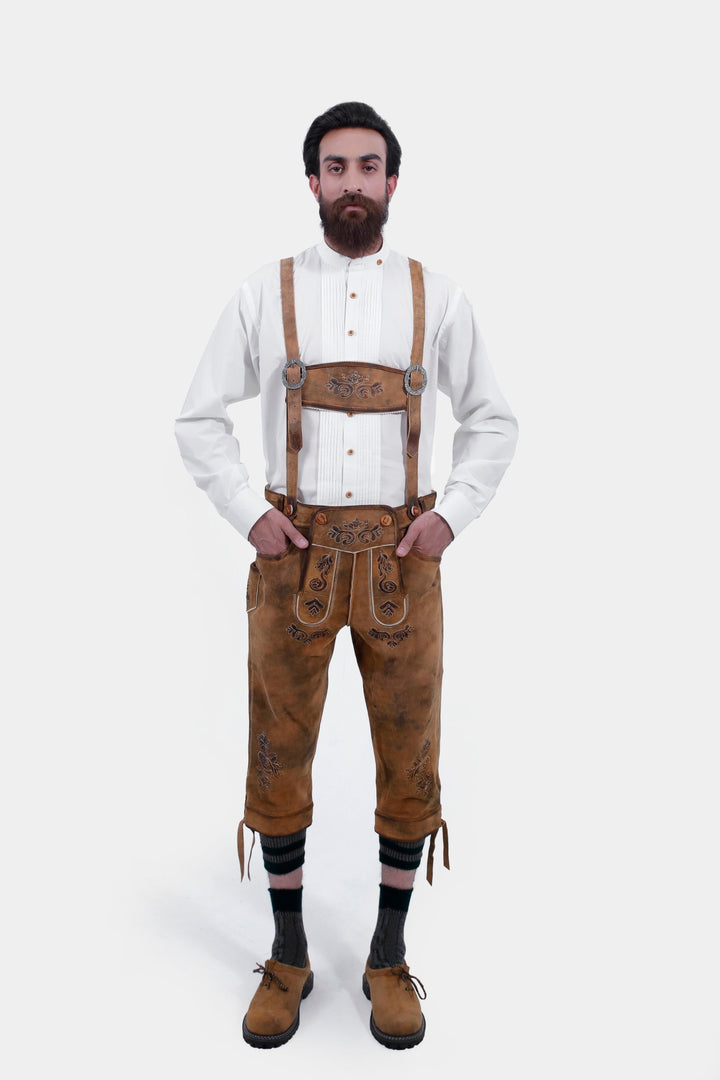 A man standing and wearing Lothar Bundhosen Outfit with suspenders. He is dressed in a white traditional shirt, brown bundhosen with intricate embroidery, dark socks, and brown shoes.