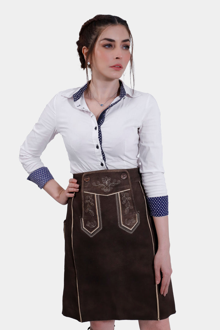 Close-up front view of a woman wearing the Louise Lederhosen Skirt in dark brown leather paired with a white button-up shirt, focusing on the skirt's embroidery, buttons, and texture.