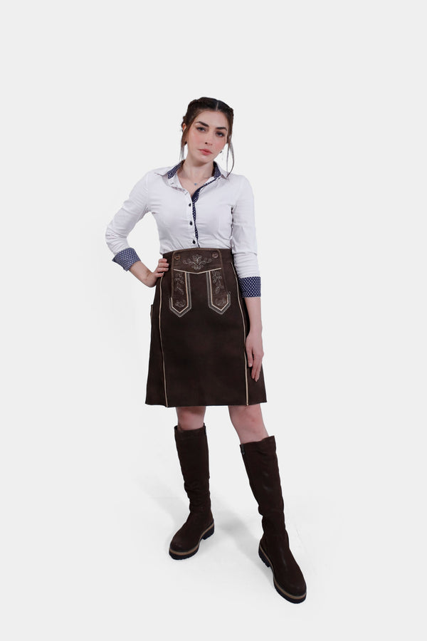 Woman wearing the Louise Lederhosen Skirt in dark brown leather paired with a white button-up shirt, standing with one hand on her hip and the other by her side.