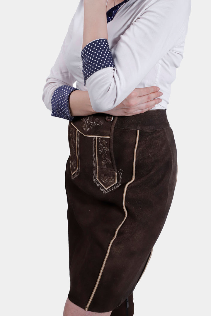 Side view of a woman wearing the Louise Lederhosen Skirt in dark brown leather, highlighting the skirt's texture, front flap with embroidery, and side pocket detail.