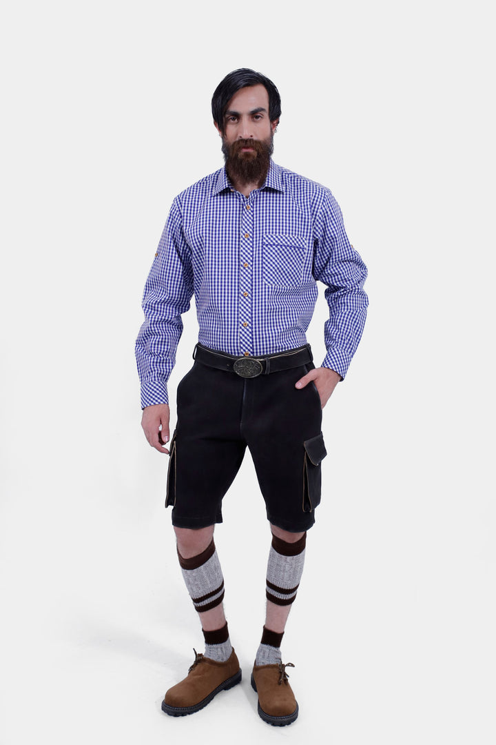 Man wearing Manor Oktoberfest Cargo Lederhosen with a blue checkered shirt, standing with hands in pockets  including  brown and grey knee-high socks and brown shoes. lederhose herren schwarz, lederhose schwarz herren​