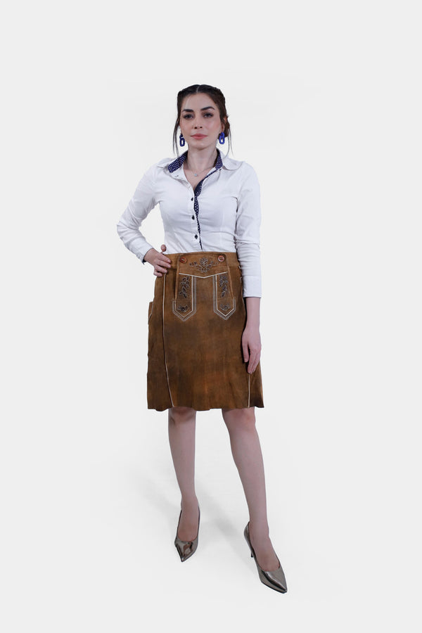 A woman wearing the Marianna Lederhosen Skirt, front view. The skirt is made of brown leather with intricate embroidery, paired with a white button-down shirt.