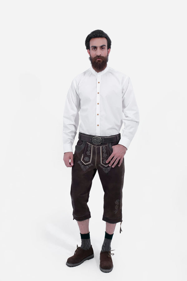 A man with a beard is wearing a white button-down shirt paired with Monte Carlo Bundhosen. The Bundhosen, made of dark brown leather, feature intricate embroidery and a decorative belt buckle. Paring with traditional socks and brown shoes 