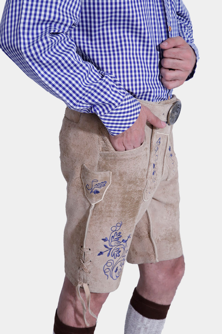 Close-up detail of the Mookie Lederhosen Outfit's blue embroidered pocket on the tan leather lederhosen, along with the blue and white checkered shirt sleeve and hand resting in the pocket.