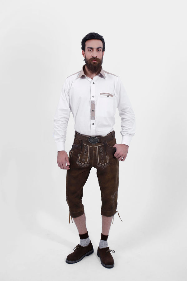 A male wearing a white shirt and Oslo Bundhosen. The Oslo Bundhosen features intricate embroidery on the front, a decorative belt buckle, and mid-length. The model's hands are by his sides, and he is wearing brown shoes with grey socks.