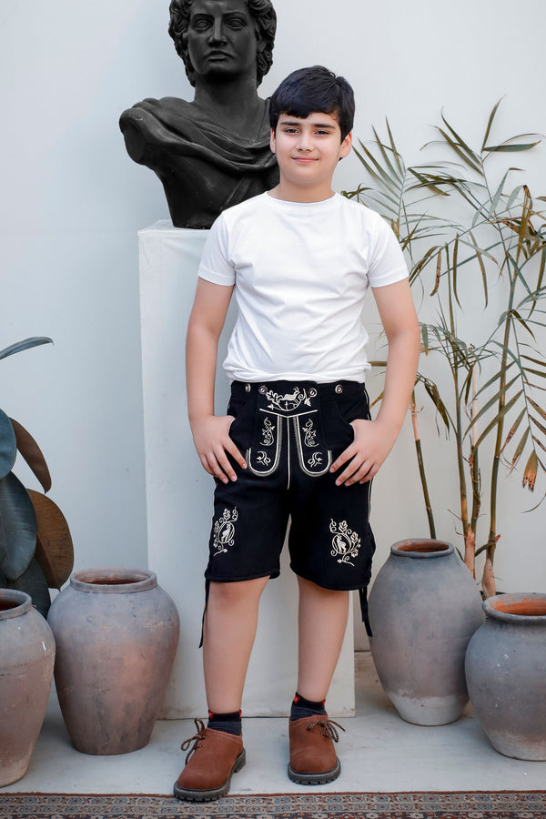 A child standing with hands in pockets, wearing Rilke Lederhosen in Midnight Edelweiss color, detailed with intricate white embroidery, and posing in front of a statue and plants.