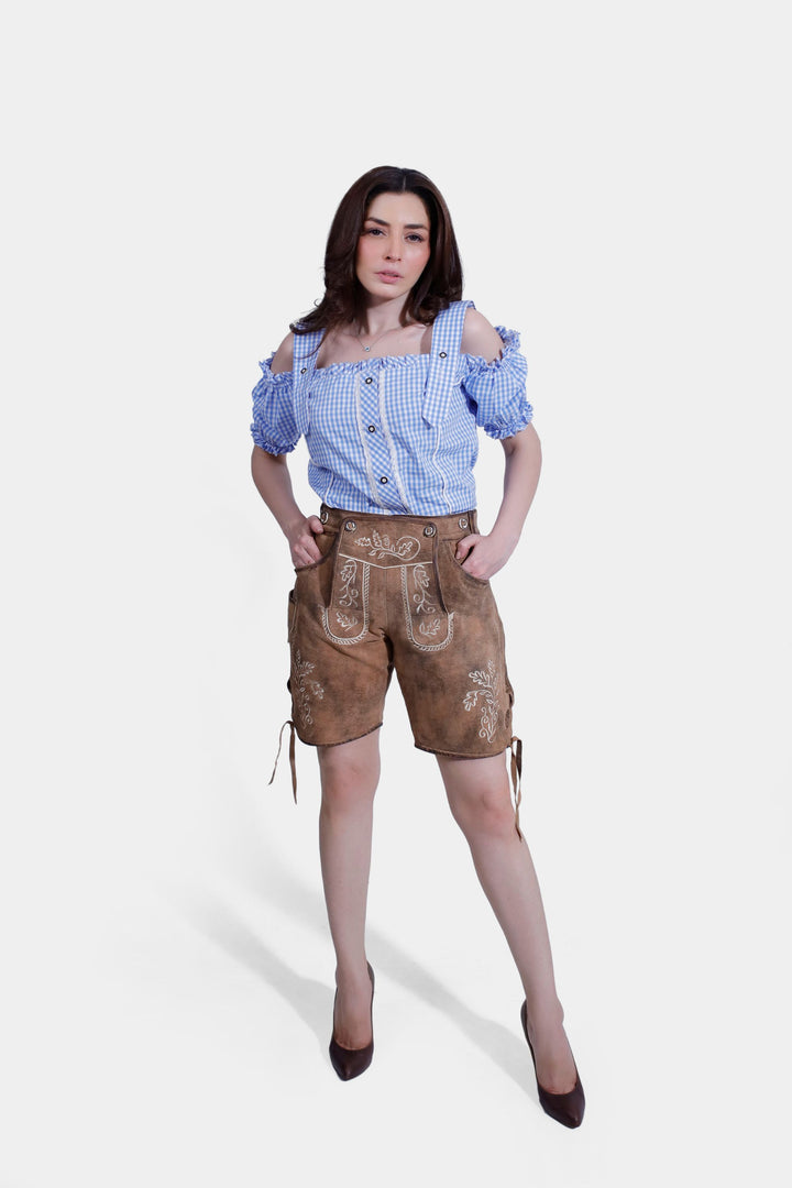 Full view of a woman wearing a blue and white checkered Bavarian shirt and light brown lederhosen with intricate embroidery, standing confidently with hands in pockets. trachten outfit damen​. trachten outfit damen​ , lederhose damen tracht​