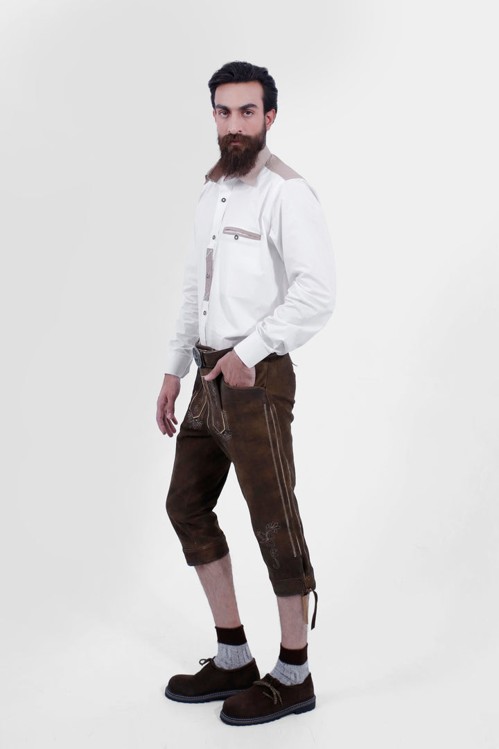 A male shown in a side profile wearing a white shirt and Schweinsteiger Bundhosen Outfit. One of his hands is in the pocket of the pants, highlighting the side embroidery and detailing, including a small side pocket. The model is wearing brown shoes with grey socks, and the mid-length pants are secured with a decorative belt.