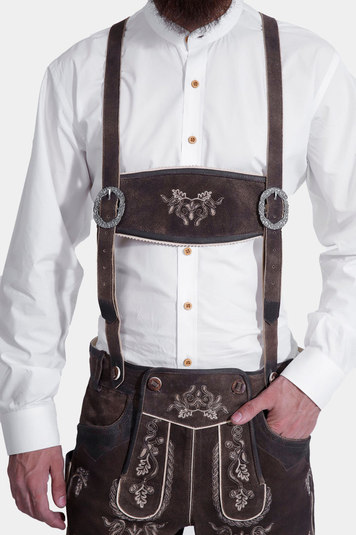 Model showcasing Spiegel Lederhosen with attached suspenders, featuring ornate buckles and embroidery on the leather shorts, along with a white shirt.