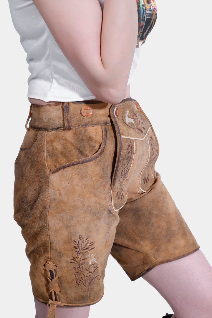 A side view of the woman in the brown leather Stefani Lederhosen, highlighting the embroidery and the belt. She is wearing a white printed blouse and has her arms crossed.