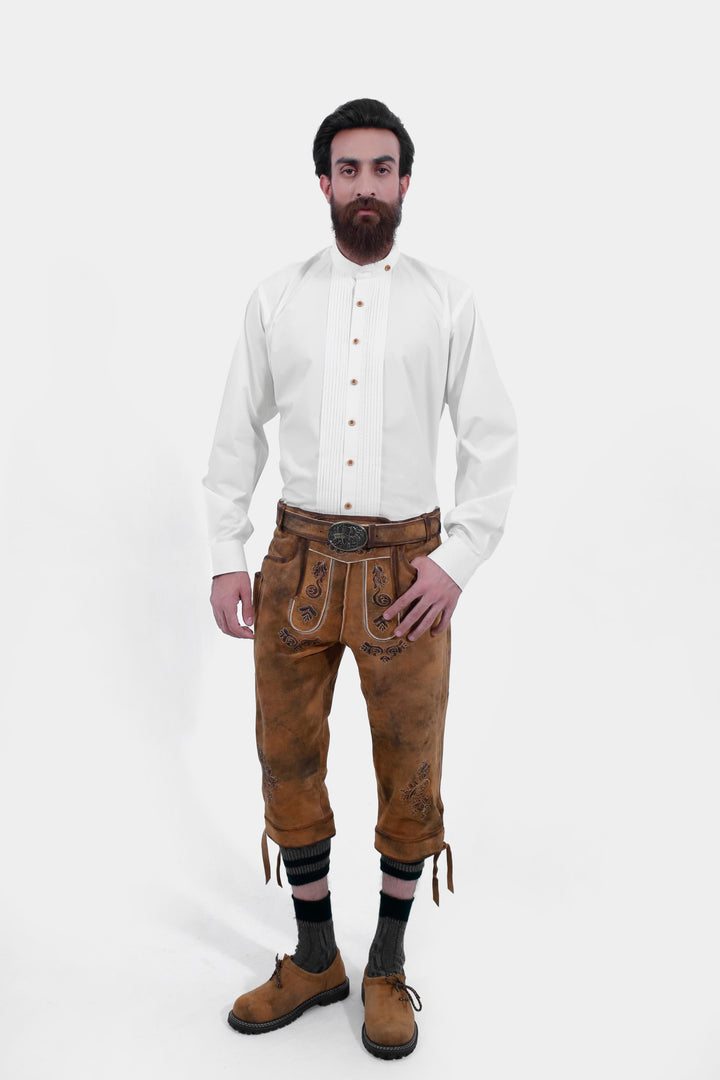 A man standing and wearing Tegernsee Bundhosen with detailed embroidery. With white traditional shirt and along with brown shoes and tan brown socks. lederhose kniebund herren hirschleder, trachten lederhose kniebund herren​