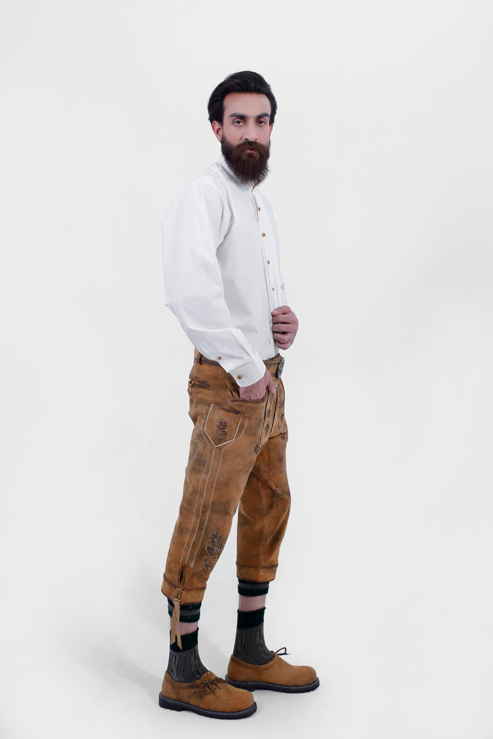 A man posing in Tegernsee Bundhosen. He is wearing a white traditional shirt, brown bundhosen with ornate embroidery, dark socks, and brown shoes, with his hand in his pocket.