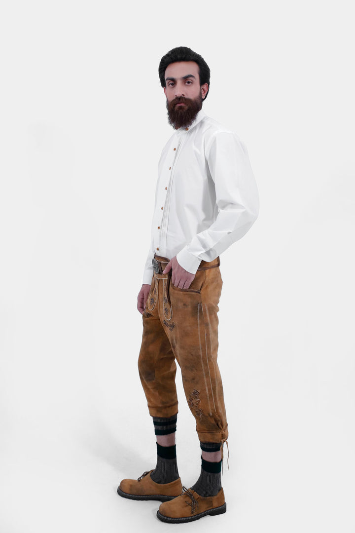 A side view of a man wearing Tegernsee Bundhosen. Wearing white traditional shirt, brown lederhosen with intricate designs, dark socks, and traditional brown shoes.