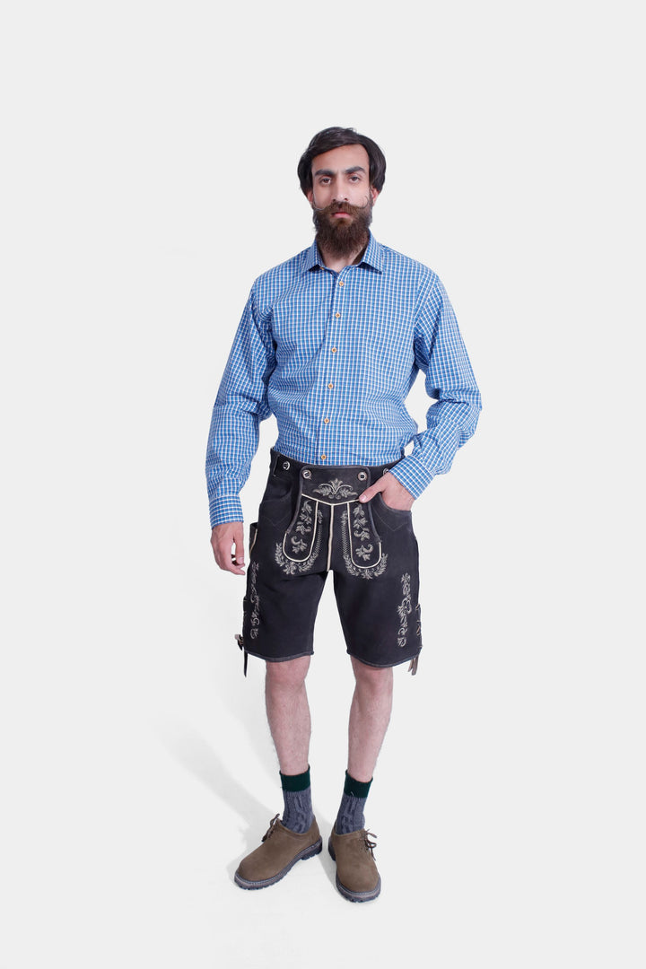 A man with a beard wearing the Timothée Lederhosen Outfit, which includes a blue checkered shirt, black leather Lederhosen with white embroidery, green knee-high socks, and brown shoes. lederhose herren tracht kurz​ ,  lederhose herren tracht​