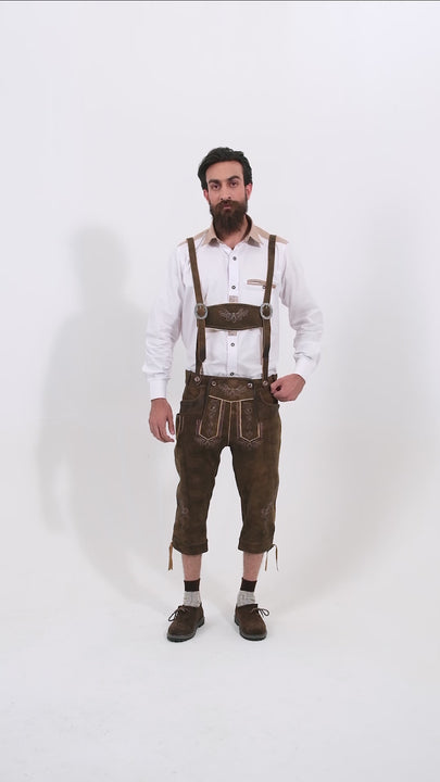 Male model wearing a white shirt and Schweinsteiger Bundhosen Outfit, featuring intricate embroidery on the front, a decorative belt buckle, and mid-length. The model's hands are by his sides, and he is wearing brown shoes with grey socks, showcasing the traditional bundhosen in a rotating video to display all angles.