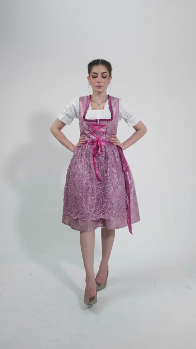 Video showcasing the Dirndl Christiane, featuring a woman gracefully modeling the outfit. She moves smoothly to display the shimmering pink lace skirt, detailed bodice with silver buttons, and satin-tied apron, highlighting the outfit's elegance and suitability for various festive occasions.