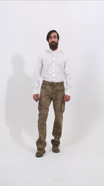 Male model wearing a white shirt and dark brown cargo lederhosen, standing with one hand in his pocket and the other by his side. The video showcases the lederhosen's multiple pockets and detailed stitching, providing a complete view of the traditional attire. The model also wears brown shoes, complementing the rustic look of the outfit as he moves to display various angles.