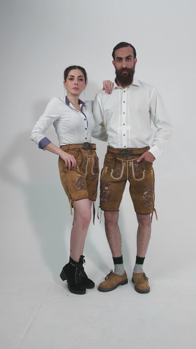 A video showcasing couple in traditional German clothing standing back-to-back. The woman is in a white blouse with blue accents and tan leather shorts with floral embroidery. The man is wearing a white button-down shirt and tan leather shorts with floral embroidery, green socks, and brown leather shoes. Both are looking serious, standing with arms by their sides.