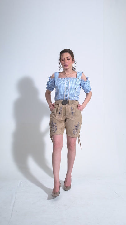 Video displaying a woman modeling beige women's Desert Sand Lederhosen, intricately embroidered in purple. She confidently walks and poses, showcasing the detailed craftsmanship of the embroidery and the fit of the lederhosen. The outfit is paired with a blue and white gingham off-shoulder top and sleek grey heels, combining traditional Bavarian style with a chic modern twist.