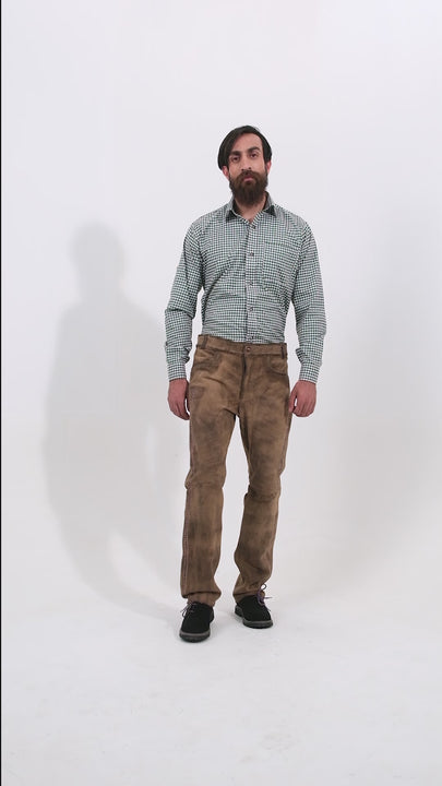 Male model wearing a green checkered shirt and beige colored Addison Oktoberfest Lederhosen, standing with one hand in his pocket and the other by his side. The video provides a comprehensive view of the lederhosen, highlighting the detailed craftsmanship and traditional design. The model is also wearing black shoes, enhancing the traditional look of the outfit as he turns to show different angles.